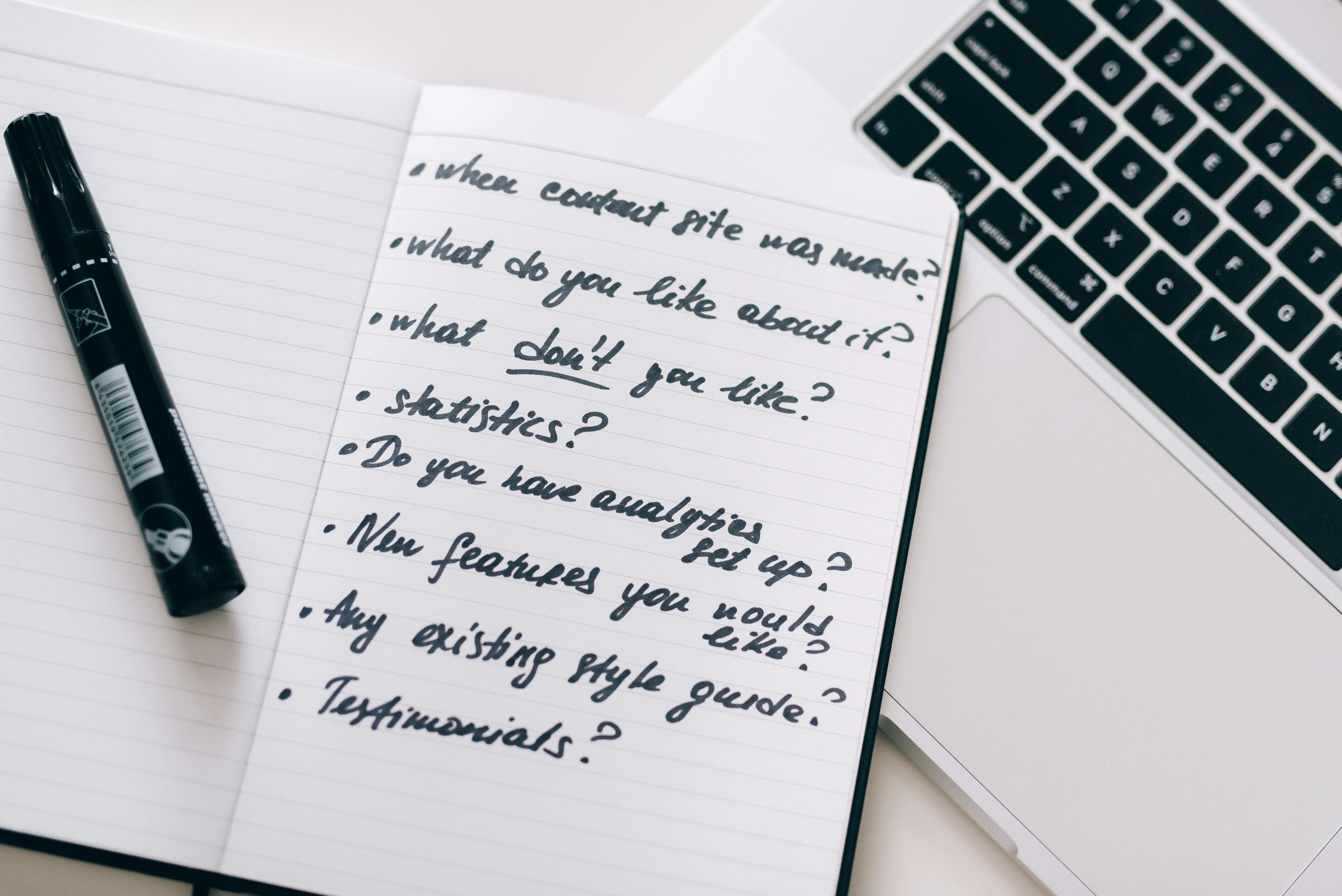 A marker placed on top of an open notebook containing handwritten notes for content strategy planning, with prompts writtent such as 'When was the content site made?', 'What do you like about it?', 'What don’t you like?', 'Statistics', 'Do you have analytics set up?', 'New features you would like?', 'Any existing style guide?', and 'Testimonials'.