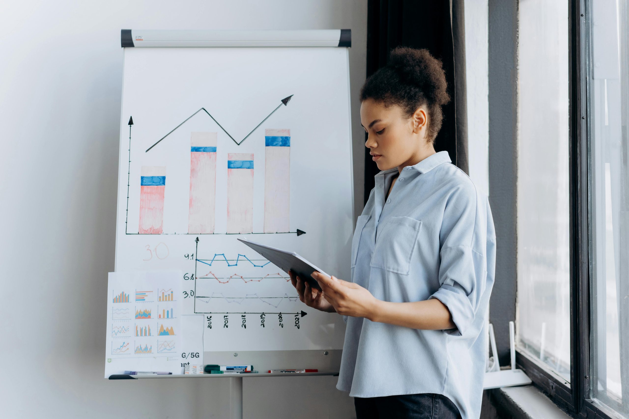 influencer marketing A woman in a semi side view reading a document on a clipboard near a glass window during an influencer marketing session, with a whiteboard in the background displaying graph results.