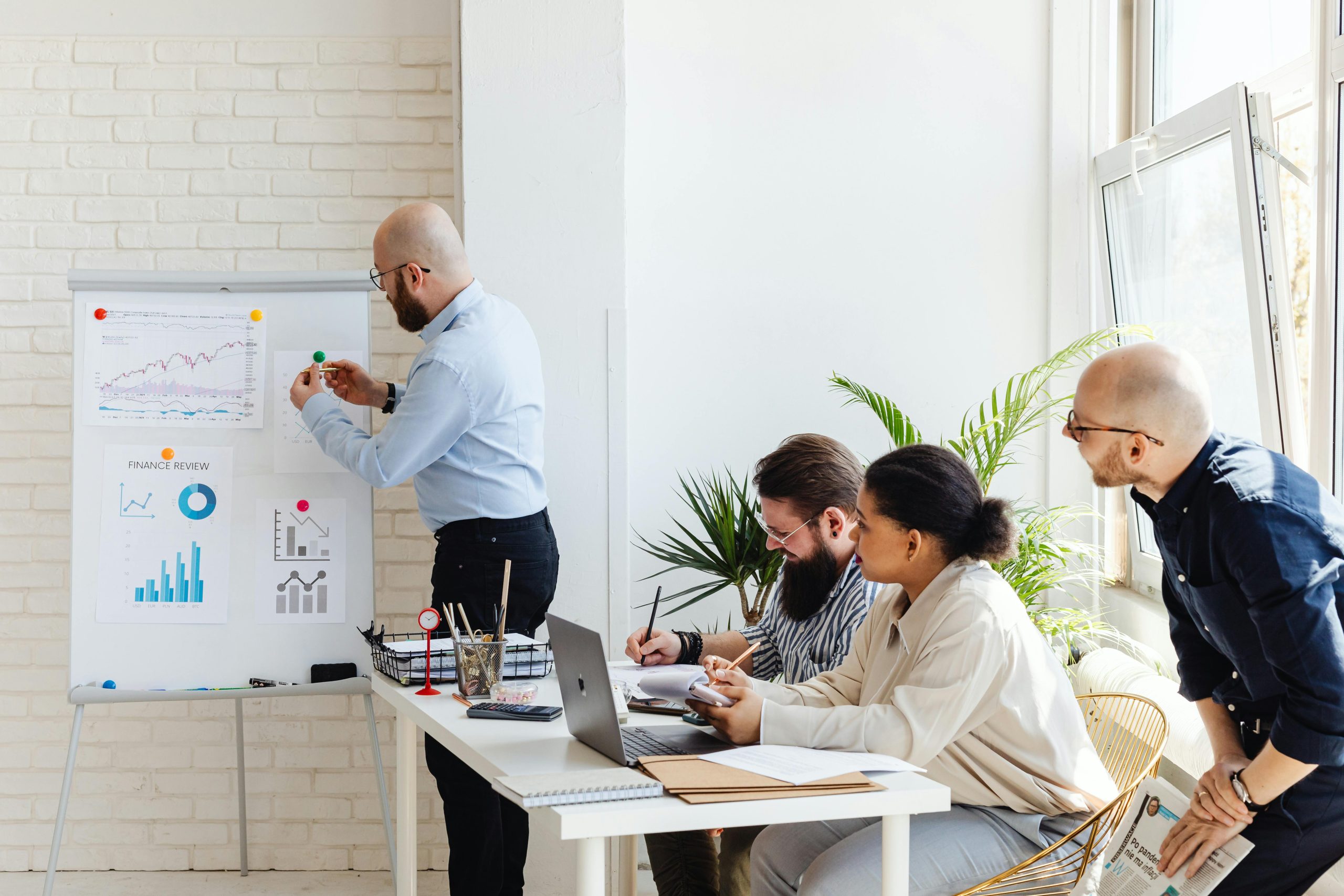 maarketing automation meeting A team leader presenting a marketing automation strategy to colleagues in a modern office, with charts and workflow diagrams displayed on a whiteboard. Two colleagues are taking notes, and one colleague is listening attentively.