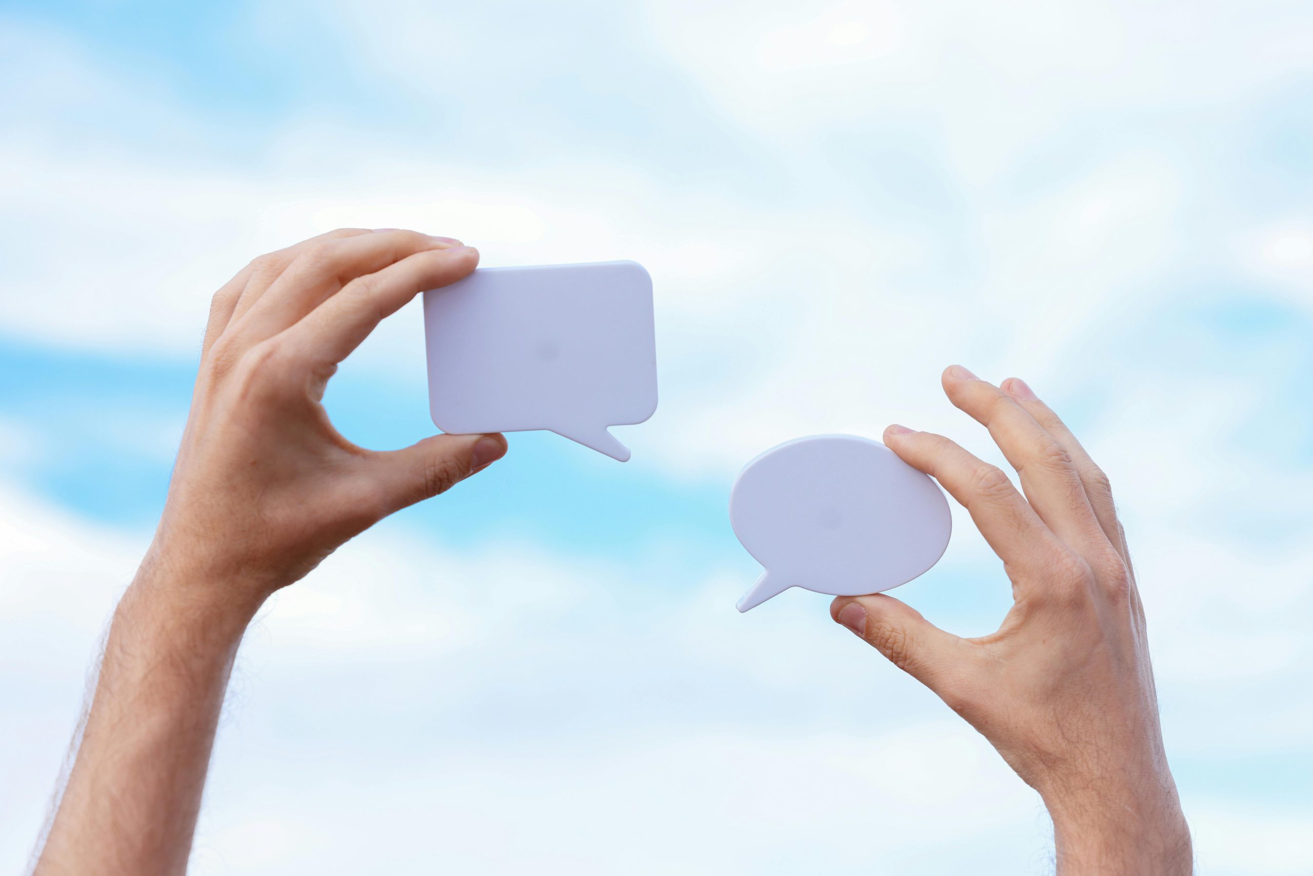 A left hand holding a paper cutout of a social media interaction icon in a box shape, while the right hand holds a round-shaped conversation bubble cutout.